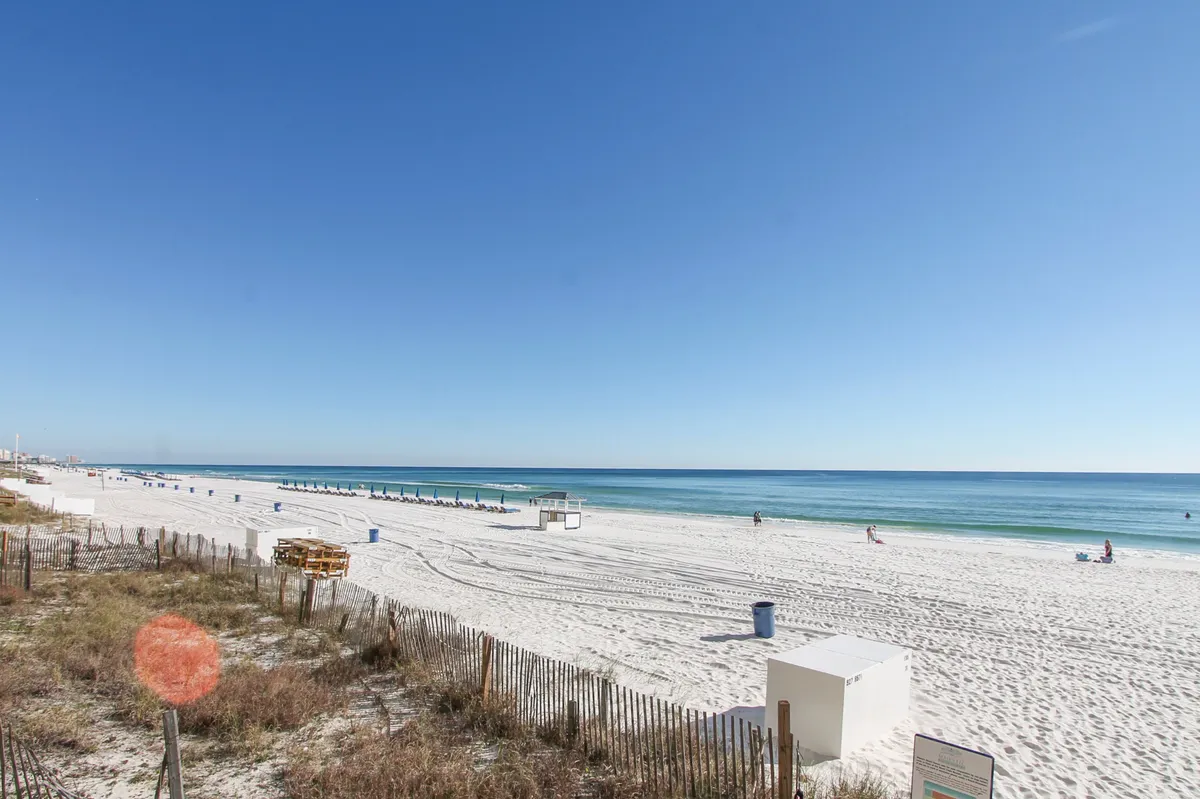 A beautiful exterior shot of a white sandy beach meeting calm blue ocean waters. Beach chairs and distant figures suggest a relaxed, social environment for sunbathing, swimming, or casual beach games.