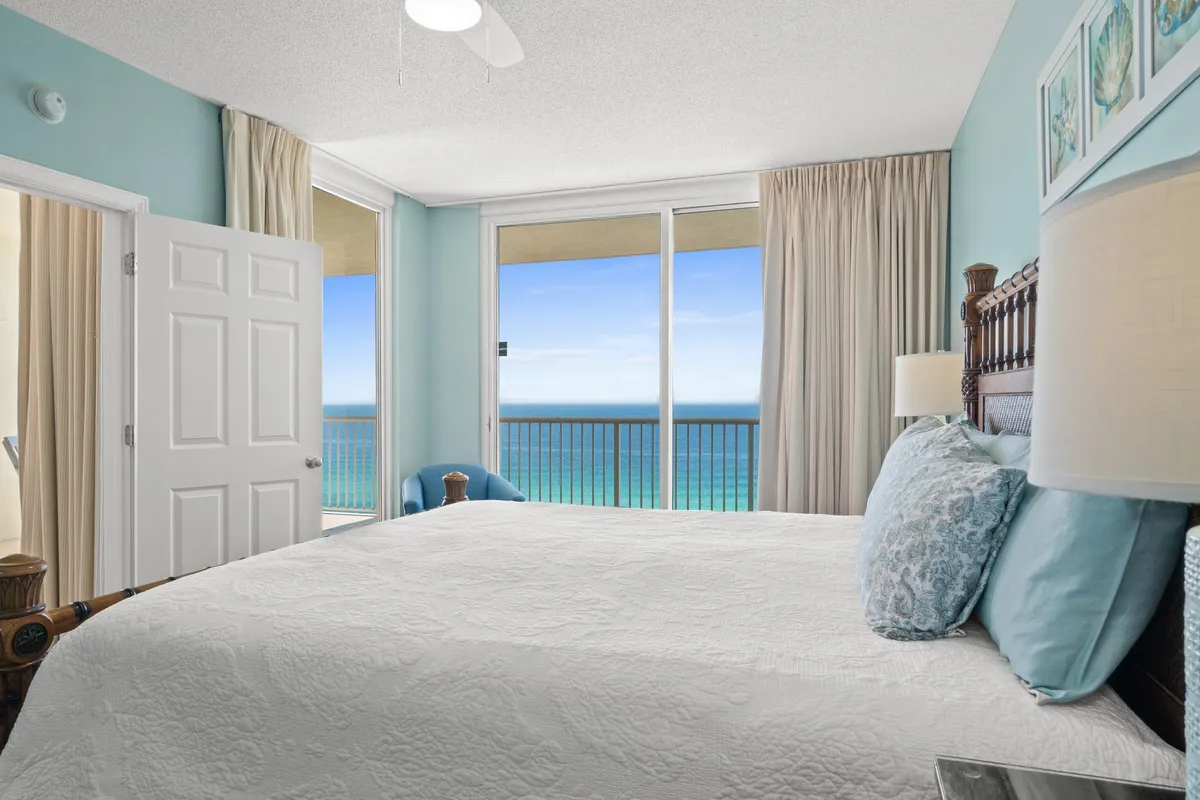 A spacious master bedroom featuring a large bed with a white quilt, blue accent pillows, and expansive windows offering a stunning ocean view. The room is filled with natural light and exudes a calm, peaceful ambiance, perfect for relaxation.
