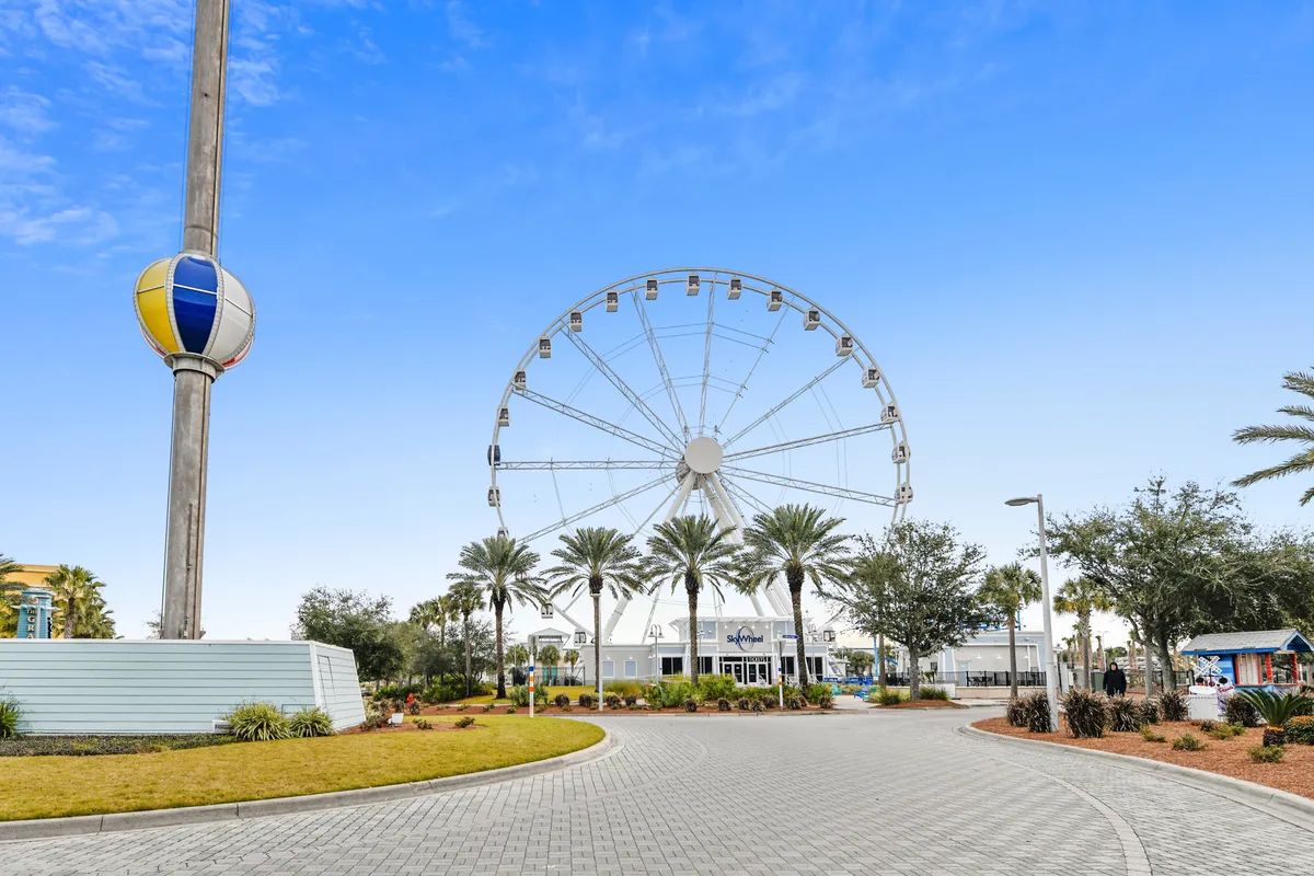 A vibrant entrance to a local attraction featuring a Ferris wheel, palm trees, and a welcoming pathway bustling with visitors. The area exudes excitement and is a hub for group adventures and memorable outings.