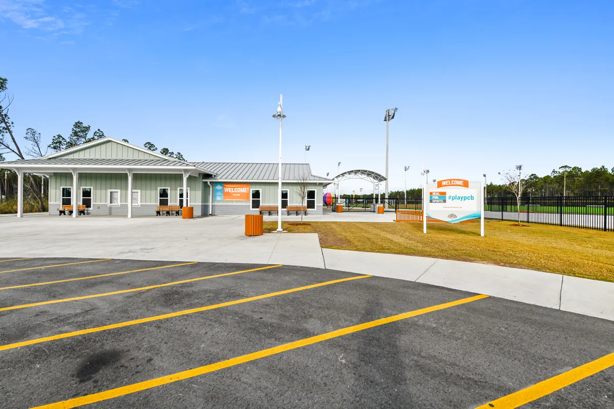 A welcoming exterior space featuring a large welcome sign, a playground area, well-manicured grass, and outdoor seating. The scene suggests a community event setting where families and children gather for play, picnics, and socializing.