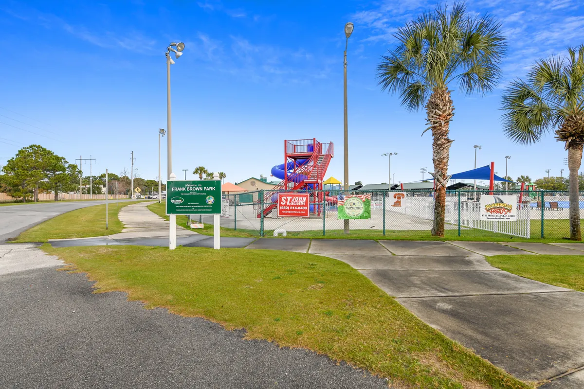 A lively exterior scene featuring a vibrant water slide, prominent park signage, palm trees, and a playground area. Families and children are engaged in swimming, playing sports, and gathering for community events, creating an energetic and joyful atmosphere.
