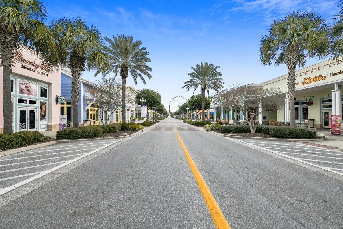 This image features a lively exterior scene with palm trees, storefronts, and the iconic Ferris wheel (Sky Wheel) visible in the distance. The promenade is bustling with people shopping, strolling, and dining.