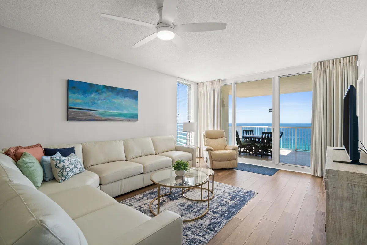 A spacious, open-concept living area with a large cream sectional sofa accented with colorful pillows. Floor-to-ceiling sliding glass doors open directly onto a balcony with sweeping ocean views, while a glass coffee table and wall-mounted TV add both comfort and style. The overall decor features bright, tranquil tones with sophisticated coastal touches.