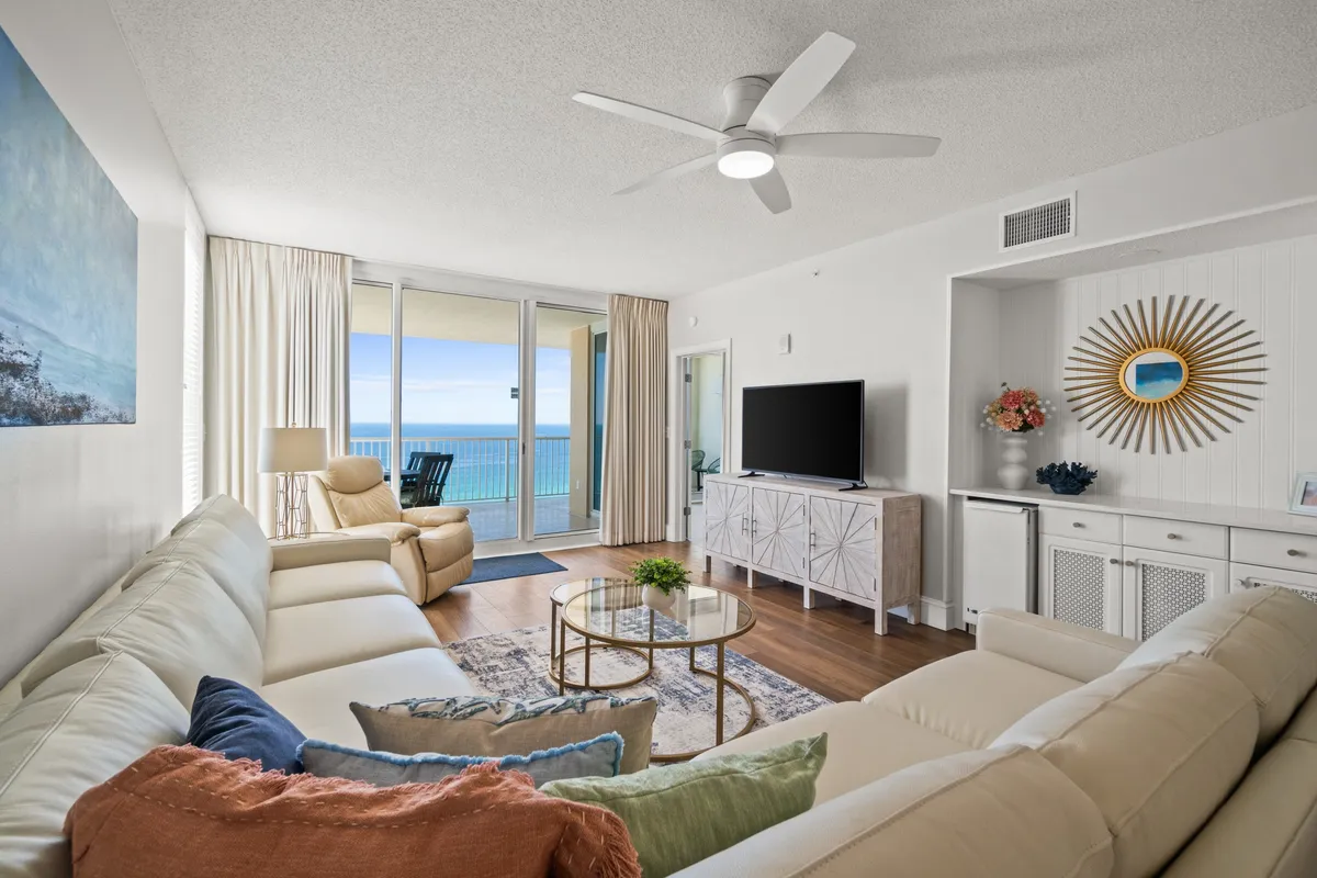 An inviting living room with a white sectional sofa adorned with colorful pillows, a glass coffee table, and a modern entertainment console with a flat screen TV. Floor-to-ceiling windows offer expansive ocean views and fill the space with natural light.