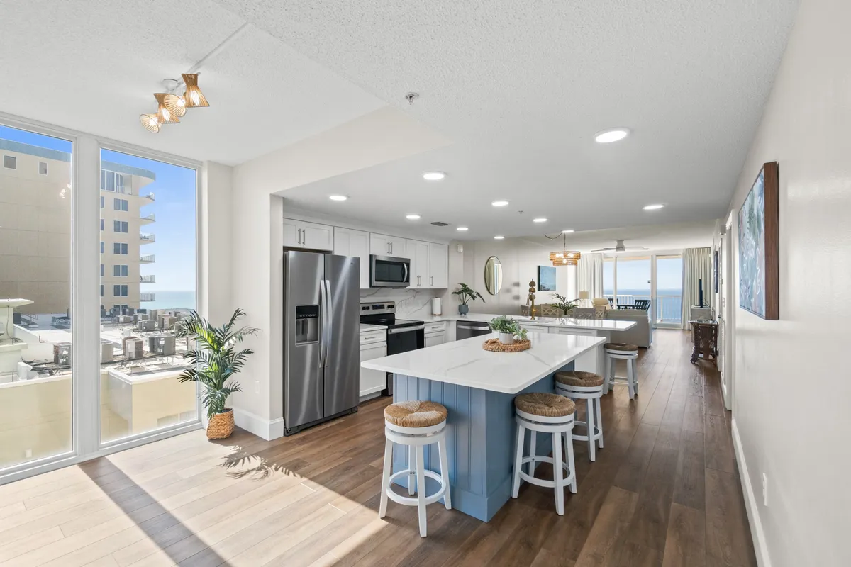 A modern, open kitchen with a large island and stools, seamlessly integrated with the living area. Floor-to-ceiling windows provide an ocean backdrop, and stainless steel appliances, along with contemporary finishes, create a welcoming and functional space for cooking and socializing. The layout highlights the open flow between kitchen and living areas.