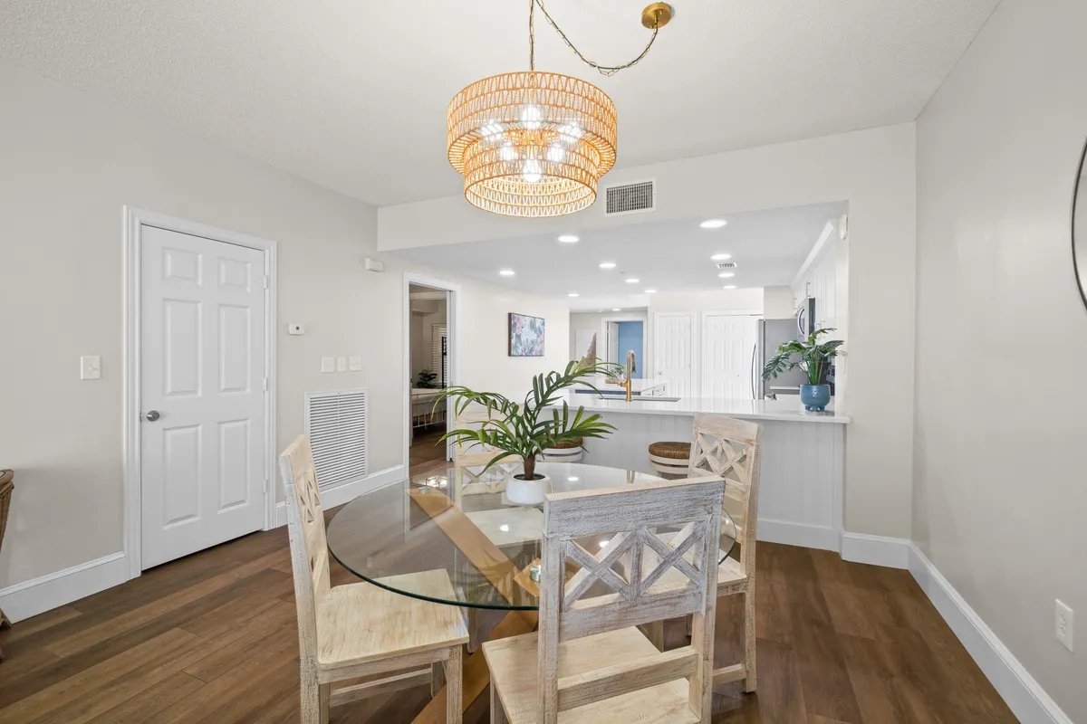 A bright and open dining area featuring a round glass table with wooden chairs, a decorative ceiling light, open kitchen with a breakfast bar and stools, and green plant accents. The layout encourages easy flow between spaces.