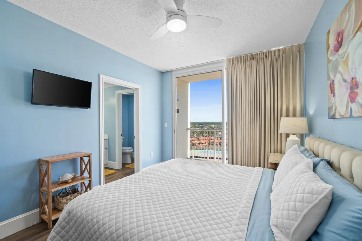 A relaxing king-sized bedroom with white quilted bedding, a wall-mounted flat screen TV, and balcony access that frames an outdoor view. An open doorway leads conveniently to the en suite bathroom, highlighting the suite's spaciousness and privacy.