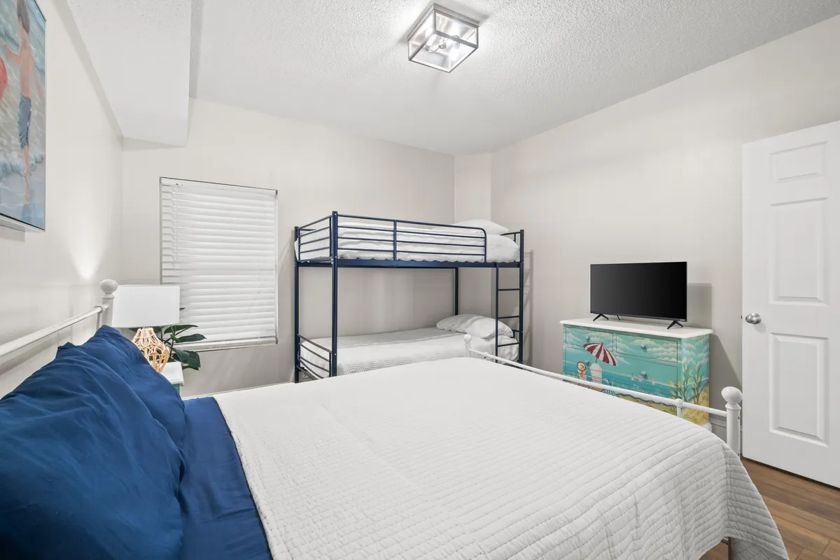 A family-friendly third bedroom featuring a white metal queen bed with navy blue pillows, a black metal bunk bed, a dresser with a beach-themed mural, and a flat screen TV. The decor is calm and inviting, ideal for families or groups of friends seeking comfort and practicality.