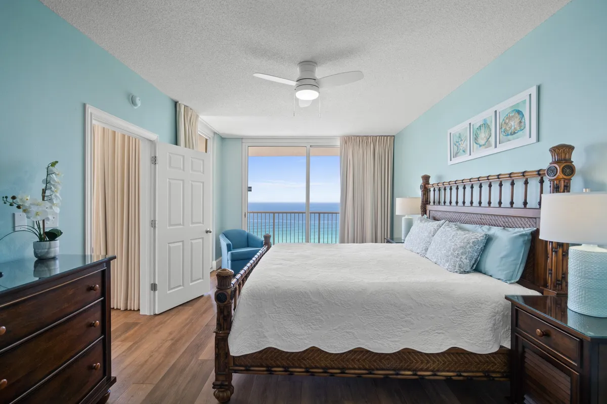 A spacious primary bedroom with a large wooden bed featuring an intricate headboard, soft blue walls, coordinated bedding, and a floor-to-ceiling balcony window offering stunning ocean views. The room includes dark wooden furniture such as a dresser and nightstand, with space to relax in an armchair and direct access to the balcony.
