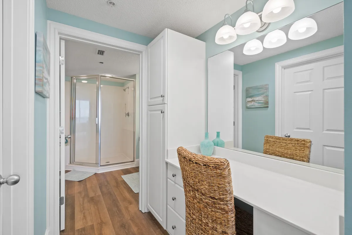 A bright, coastal-inspired bathroom with a large mirror above the vanity, a woven chair, glass shower enclosure, and white cabinetry. The space is inviting and functional, designed for personal grooming and relaxation after a day at the beach.