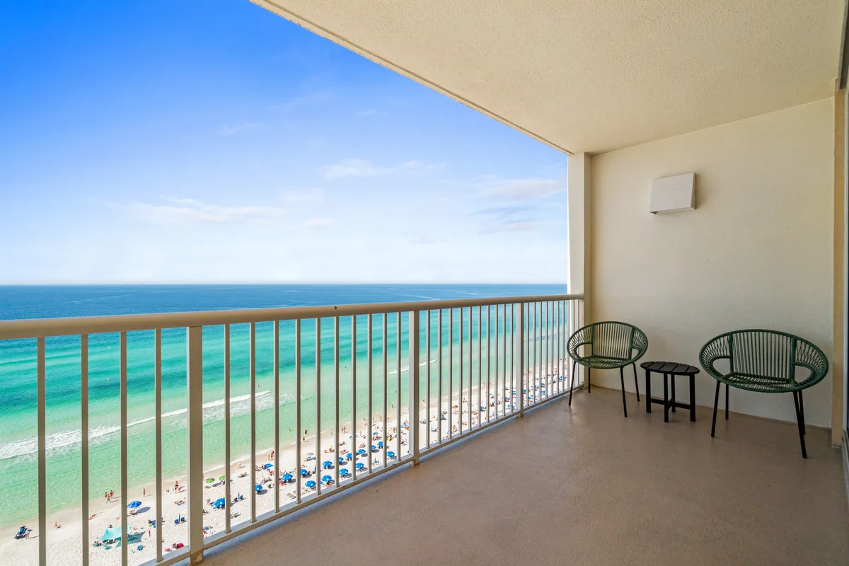 A massive, furnished private balcony overlooks the turquoise Gulf waters and white sandy beach below. Minimalist outdoor chairs and a table invite guests to relax and take in panoramic views. Beach umbrellas and people enjoying the shore emphasize the vacation atmosphere and the balcony's role as a social gathering spot.