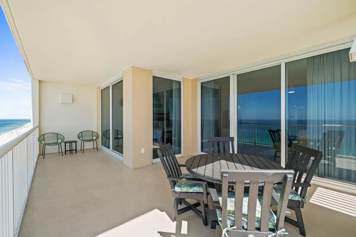 A round dining table with chairs sits on a spacious balcony, framed by floor-to-ceiling sliding glass doors. The balcony overlooks a stunning beach and ocean view, embodying the indoor-outdoor living highlighted in the content.