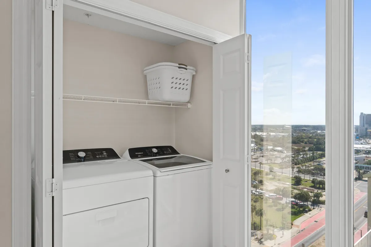 A bright laundry area featuring a modern washer and dryer set, a laundry basket on a shelf above, and open closet doors. Natural light from a window helps create an efficient, home-like feel.
