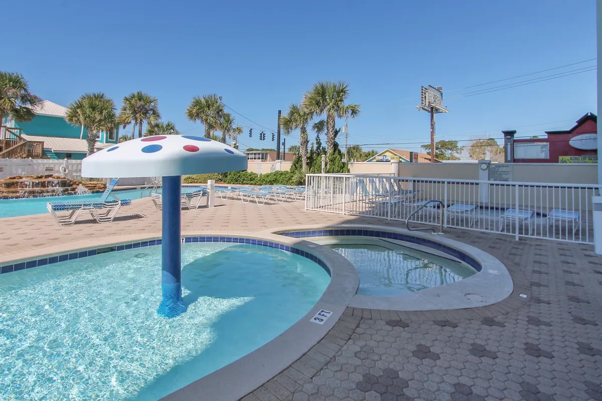 A cheerful children’s splash pool with a mushroom fountain is situated next to a small hot tub, surrounded by lounge chairs and palm trees. The space is lively yet safe, suggesting a family-friendly environment with options for both kids and adults to enjoy water activities or relax.