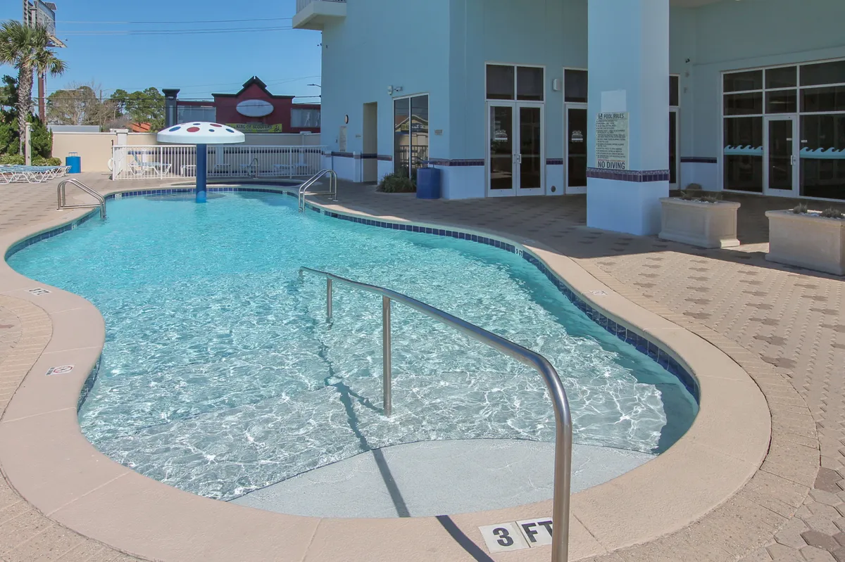 A curved, sparkling swimming pool sits at the resort's entrance, surrounded by a poolside deck with a mushroom water feature. Sun loungers, glass doors, and the inviting pool area highlight the resort's amenities for guests to enjoy together.