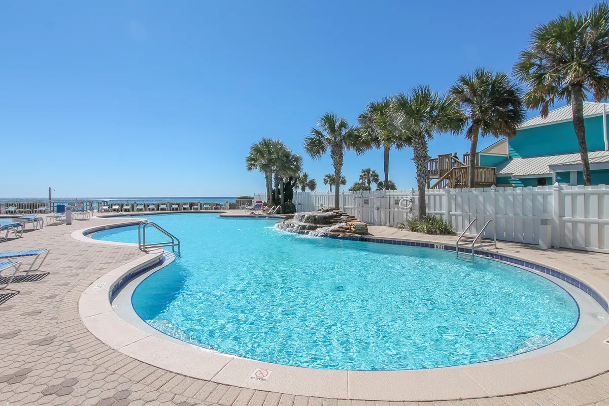 An inviting outdoor swimming pool with a waterfall feature is framed by palm trees, poolside lounge chairs, and the ocean visible just beyond the pool area. The scene suggests a relaxing resort environment where guests can swim, sunbathe, or socialize with the soothing backdrop of water and coastal landscapes.