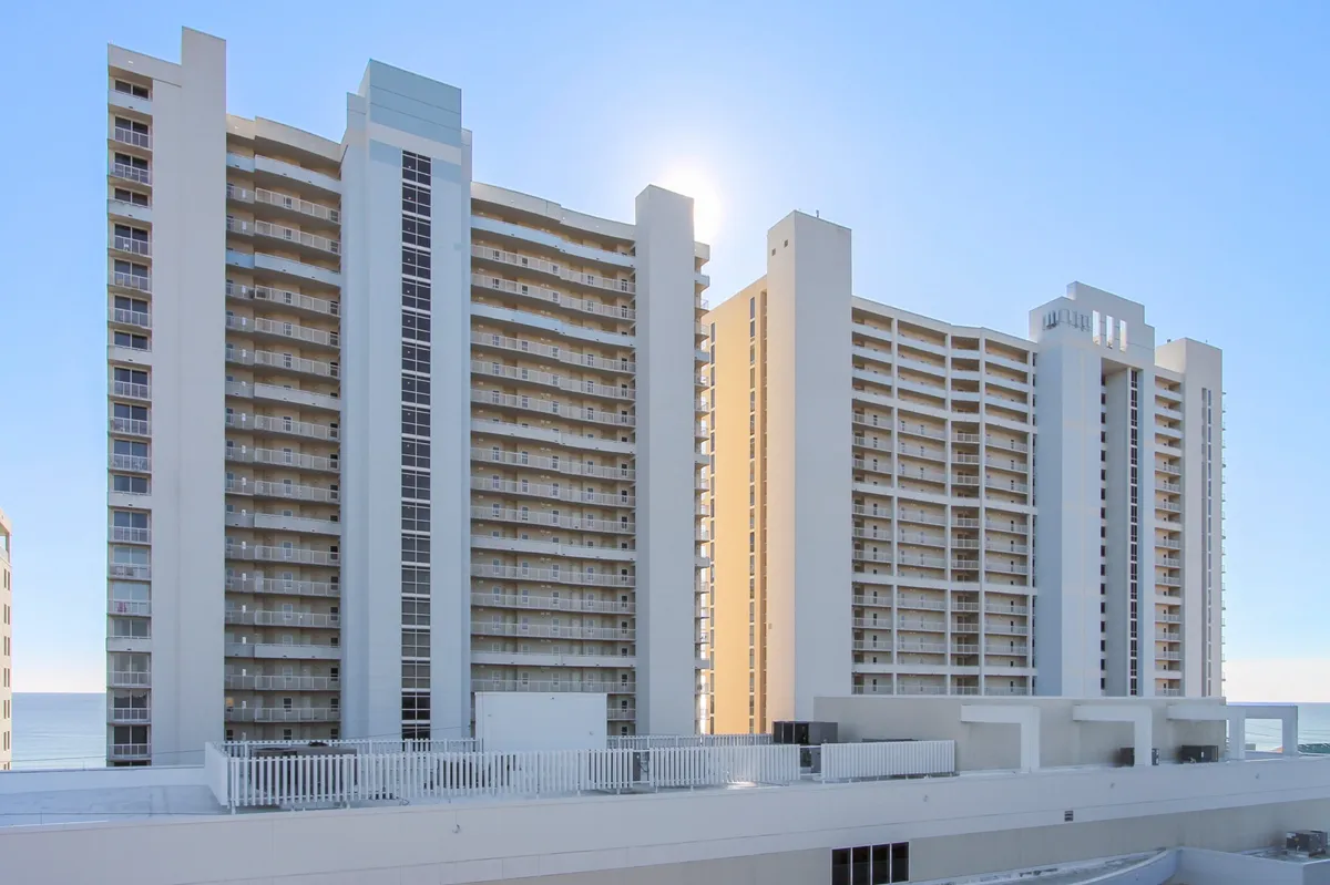 This image captures a tall white condominium building, likely Majestic Beach Resort, with multiple balconies overlooking a bright blue sky and a glimpse of the ocean. Sunlight reflects off the building, emphasizing its height and luxurious vibe.