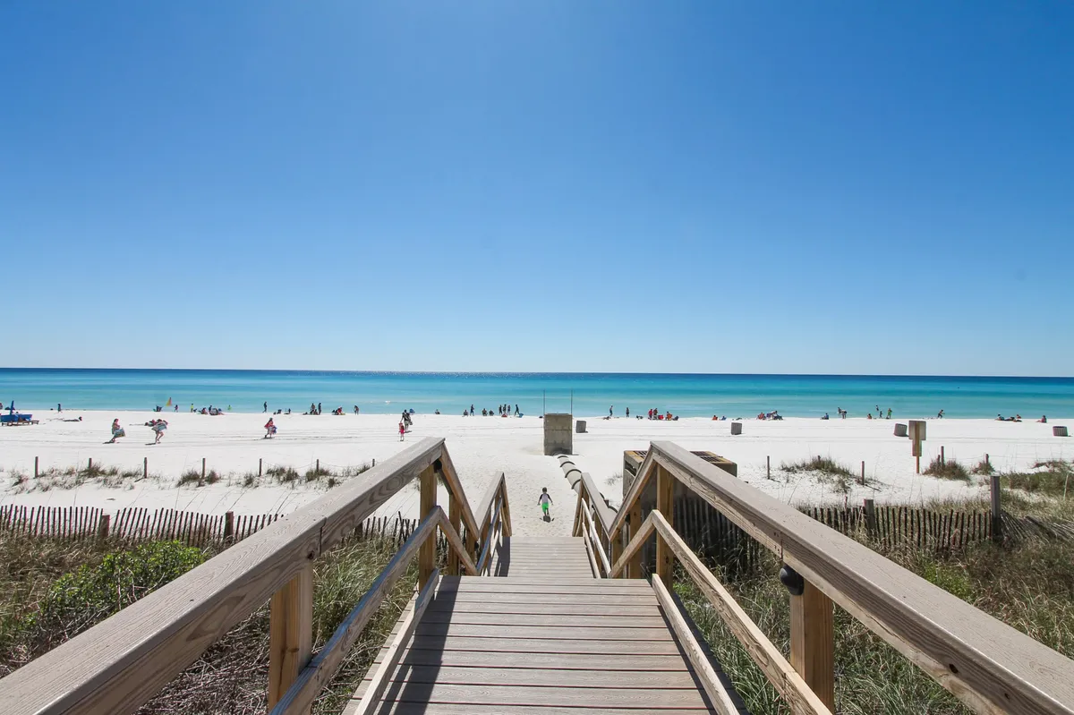 A wooden boardwalk leads directly to a pristine white sandy beach and turquoise gulf water, with people enjoying the shore in the distance. The scene encapsulates easy, private access to the beach and the seamless transition from resort amenities to natural beauty.