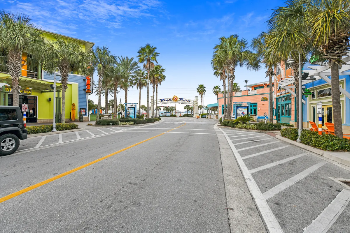 A cheerful street scene lined with colorful storefronts and palm trees, with a welcoming plaza entrance sign. The clear blue sky and inviting streetscape communicate the convenience of walking to local shops and restaurants just steps away from the resort.