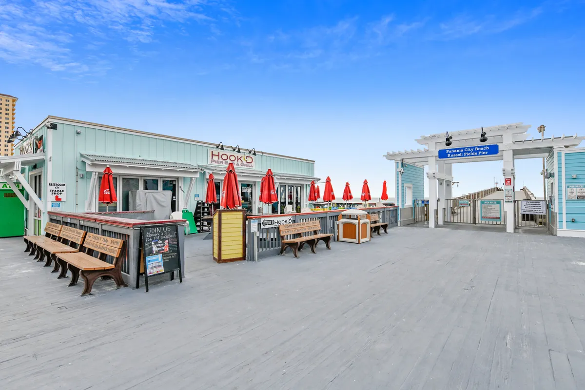 The Hook’d Pier Bar & Grill and the entrance to Panama City Beach’s Russell Fields Pier create a vibrant coastal scene, with red umbrellas, benches, and the ocean beyond. The atmosphere is lively and inviting, ideal for exploring local dining and attractions.