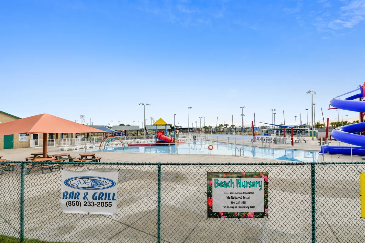 A vibrant children's splash pool area featuring colorful water slides, interactive play structures, and shaded picnic tables with umbrellas. The pool is safely fenced and surrounded by clear safety signage, creating a playful and secure environment for families with young children.